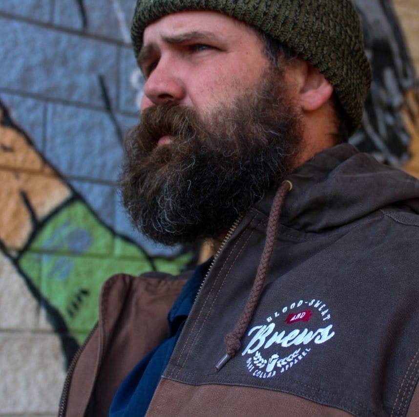 Bearded man in brown hoodie with "Brews and Battle Stories" logo, outdoor urban scene, graffiti background, cold weather attire, rugged masculine appearance, adventure, storytelling, brewing culture, community engagement, beer enthusiast, outdoor lifestyles.
