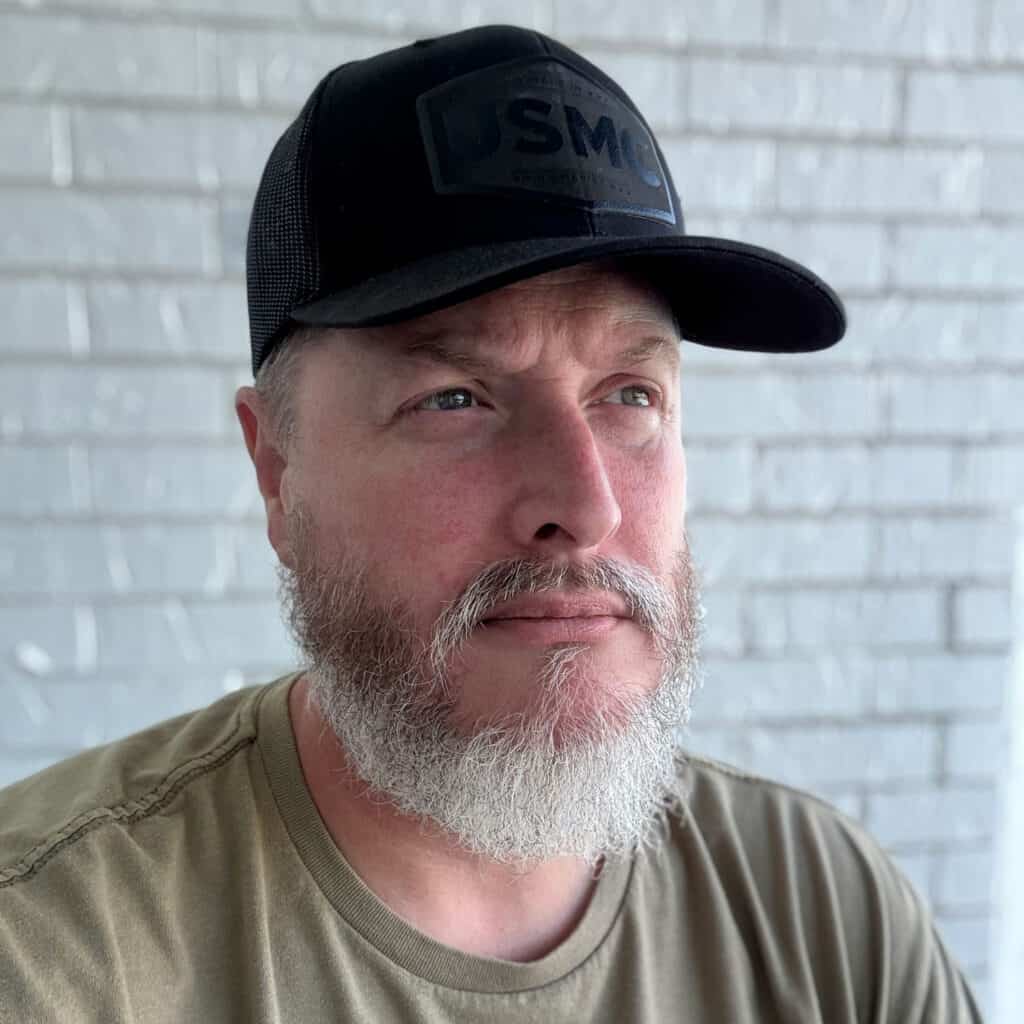 Bearded man wearing a black cap with "USMC" patch, looking thoughtfully into the distance against a white brick wall background.